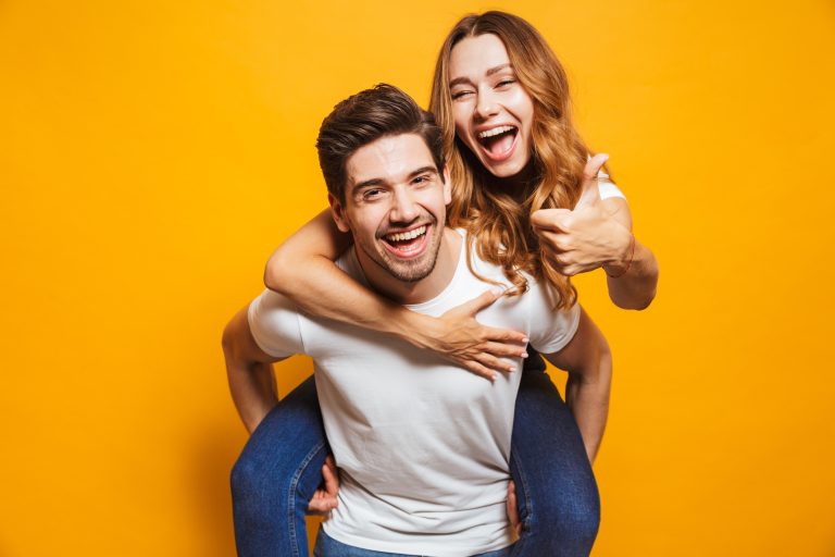 Man smiling while giving a piggyback to a happy woman giving a thumbs up