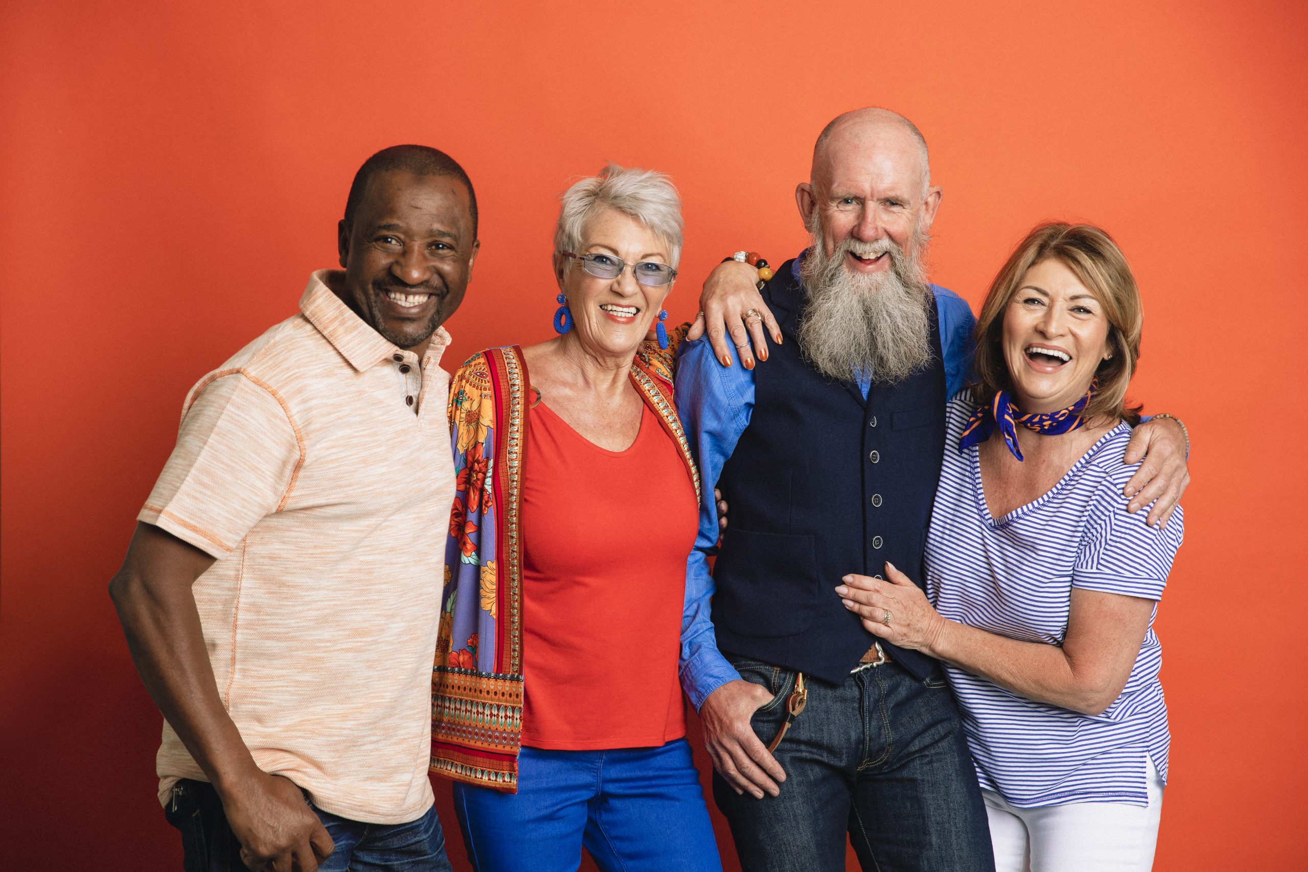 A group of older friends laughing together with their arms around each other standing in front of an orange studio background looking at the camera.